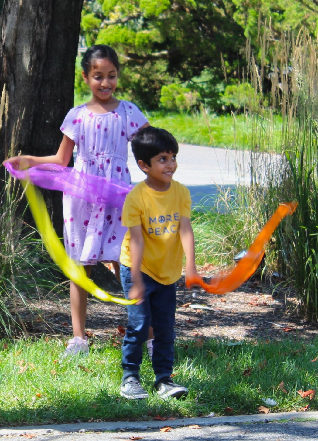 Two smiling children standing in the grass and holding purple, yellow, and orange scarves.