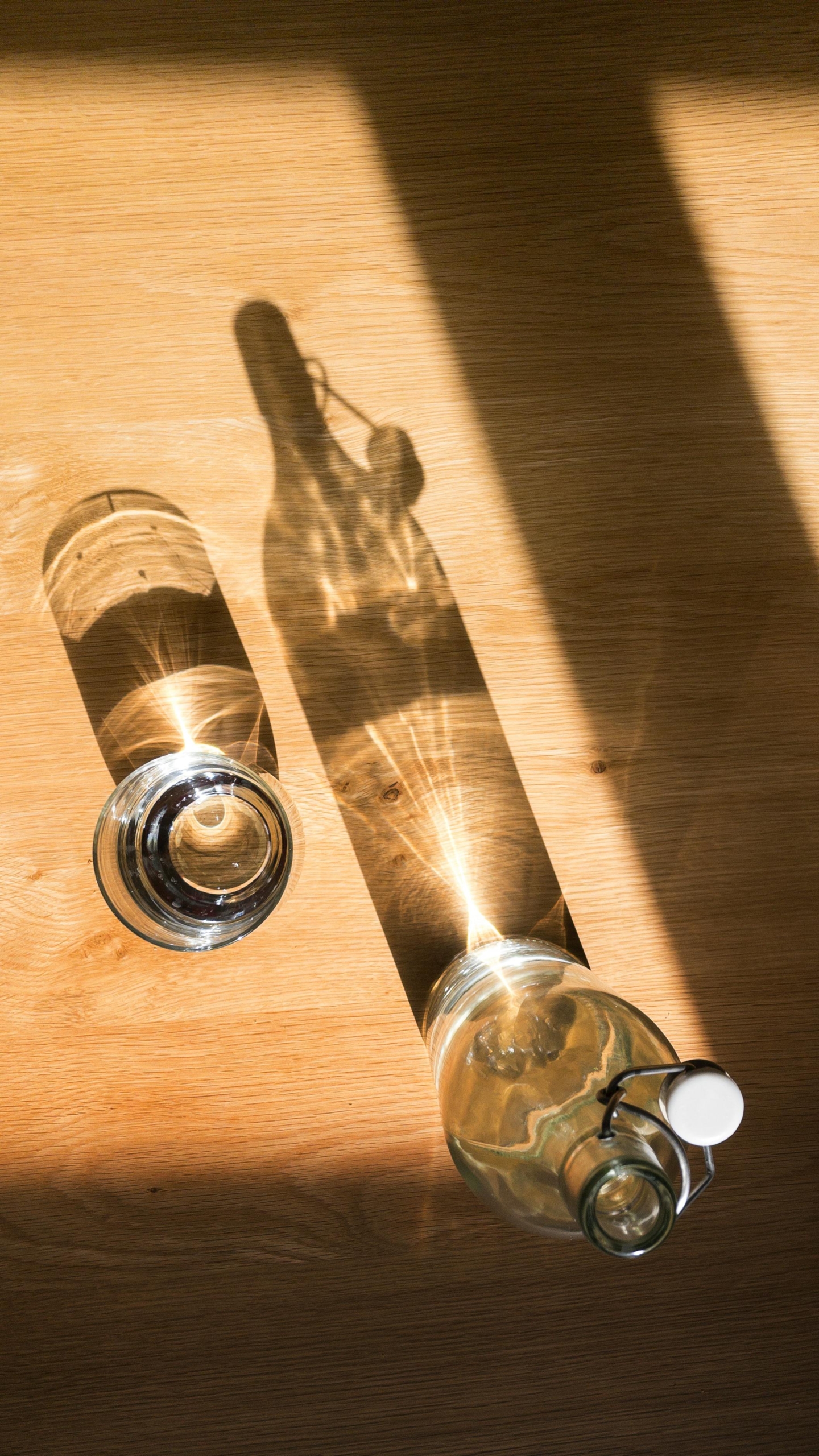 A glass bottle and drinking glass cast a shadow on a sunny wooden table.