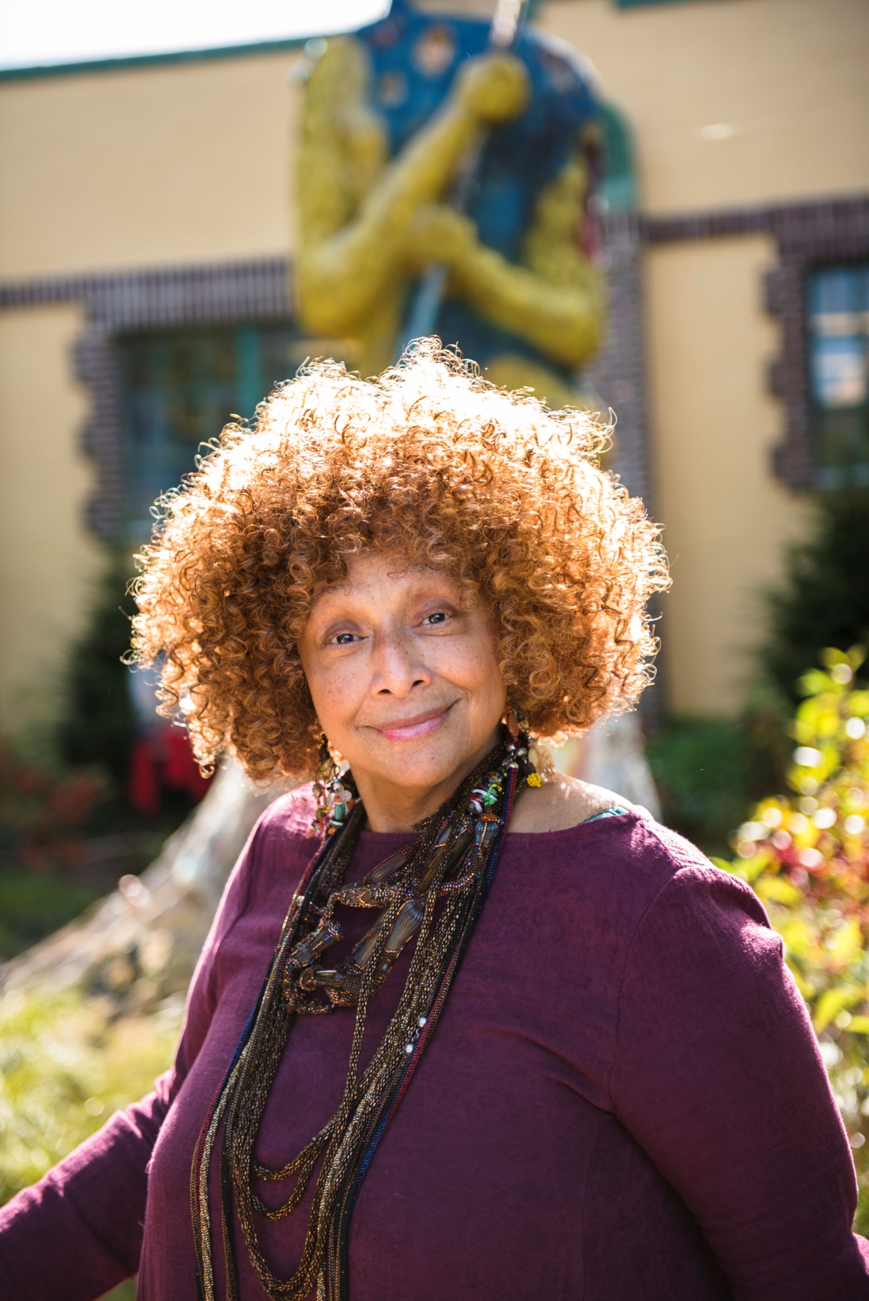 Joyce J. Scott in the Water Garden at Grounds For Sculpture. She is smiling and wearing a purple shirt and beaded necklaces.