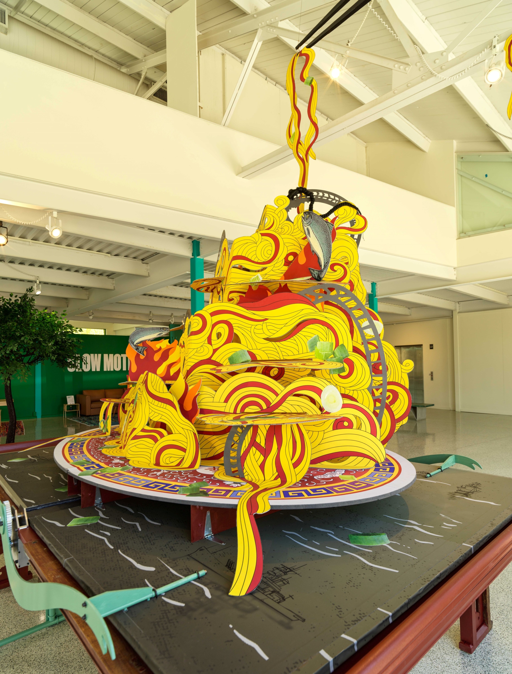 A side view of 'Noodle Mountain,' a large pop-up book of yellow noodles with green onion and salmon details.