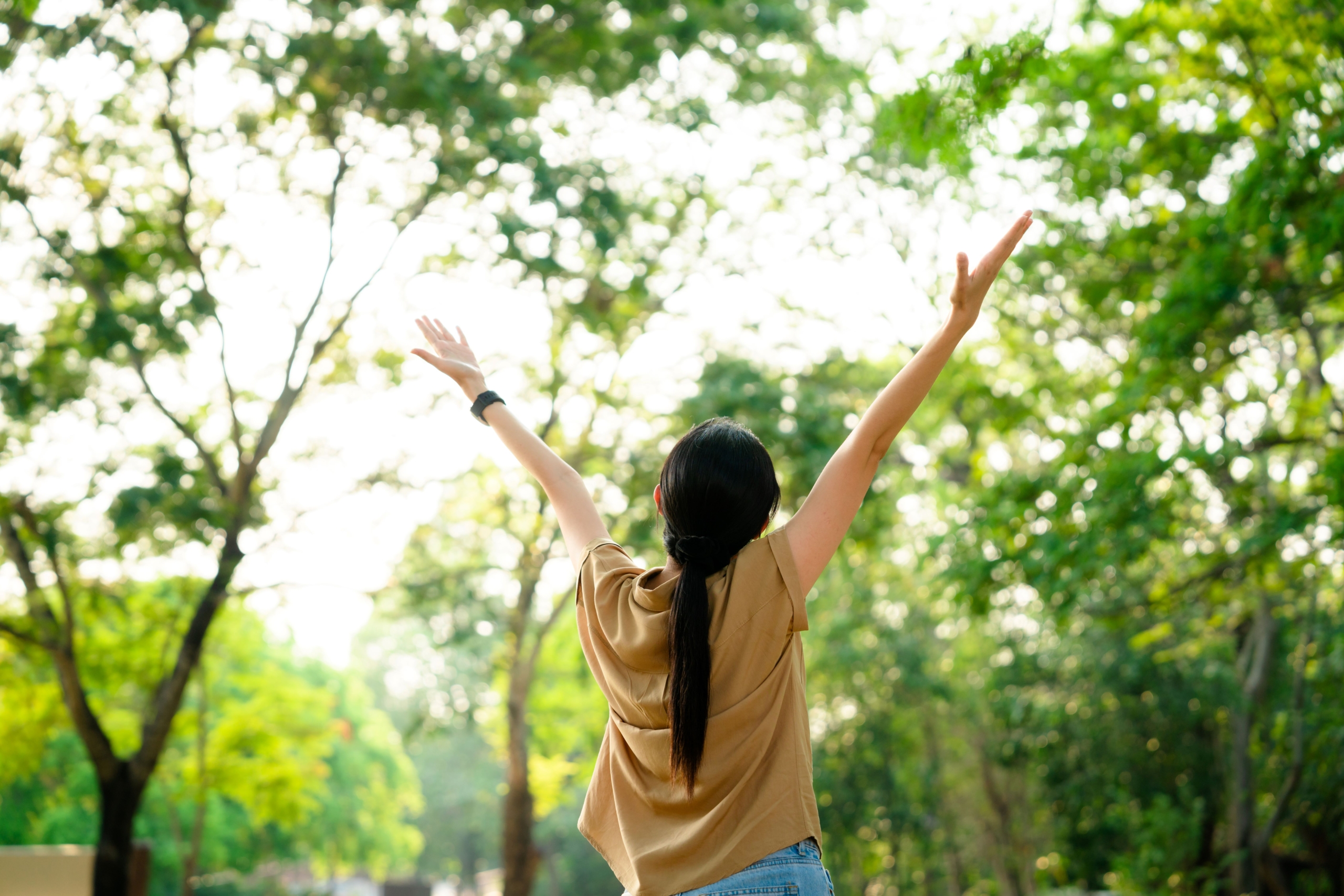 A woman in a yellow shirt holds her arms up, facing away from the camera, in a forest.