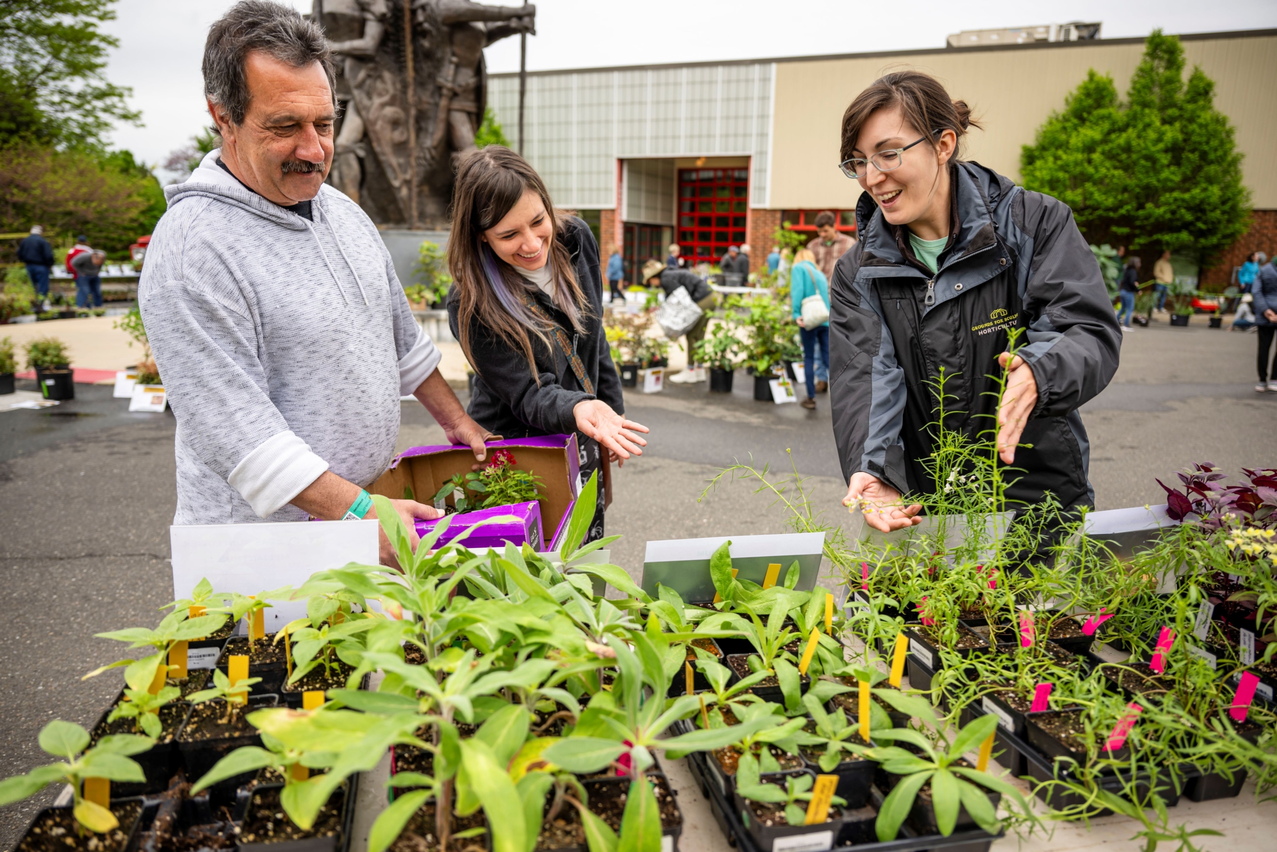 A staff member showing two guests a table of potted plants outdoors.