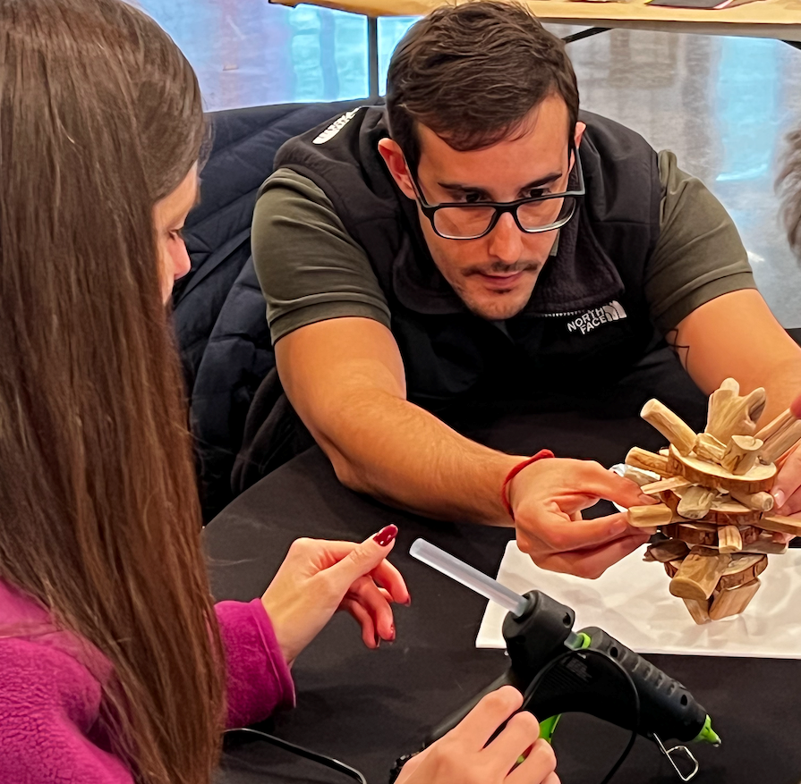 Two people working together to build a small wooden sculpture on a table.