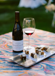 An outdoor shot of a glass of red wine, the red wine bottle, and a plate of Wagyu sliders on a table.