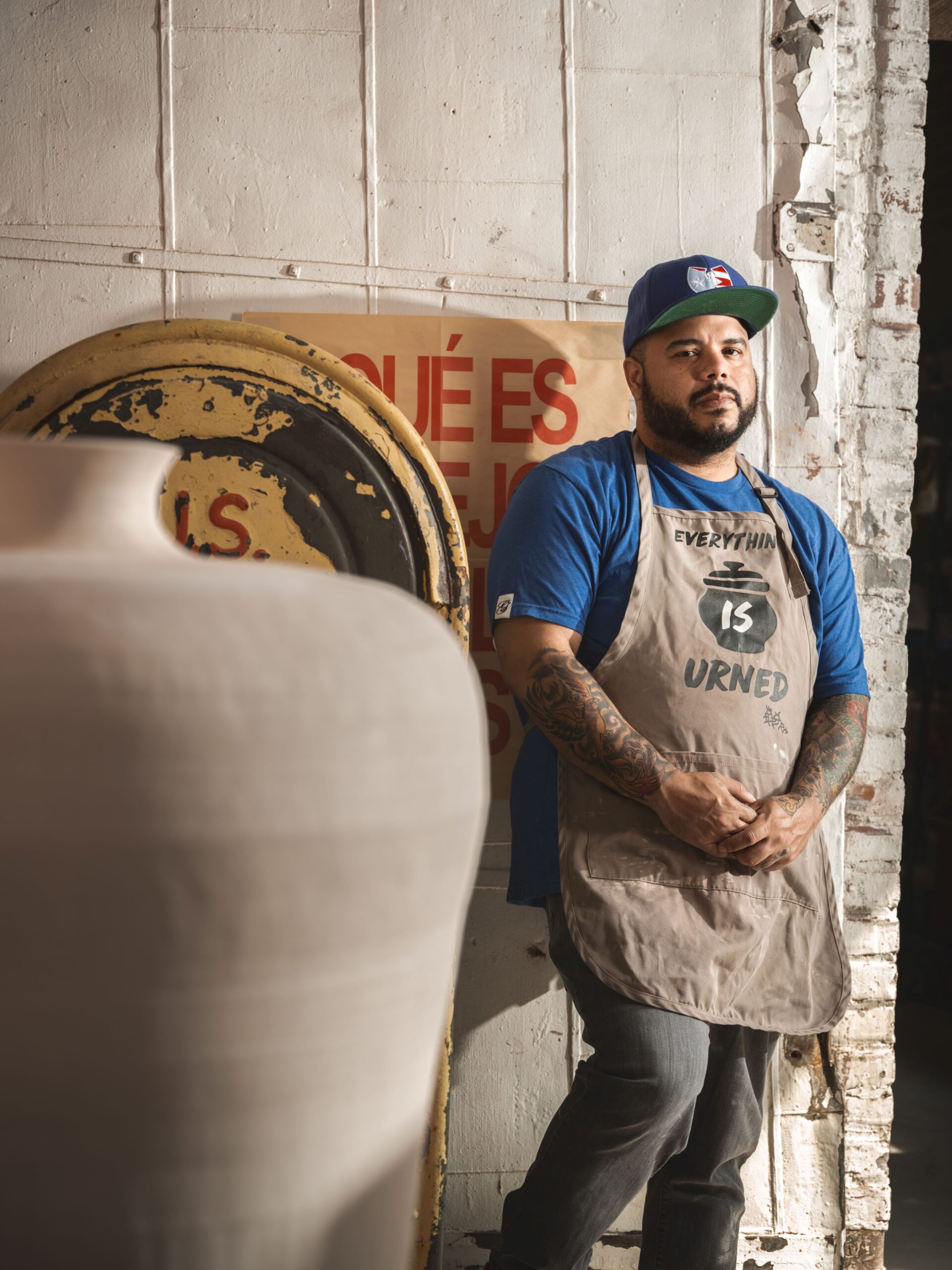 Roberto Lugo in a baseball hat and a tan apron standing next to a large clay pot.