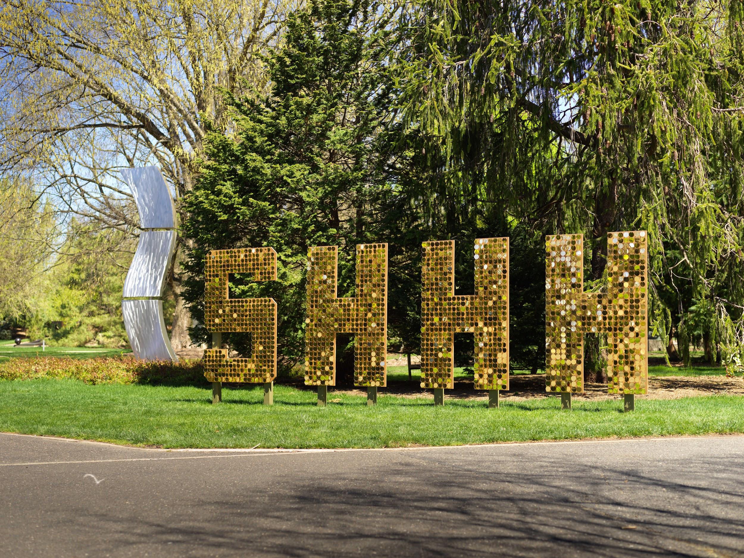 Seven feet tall letters with sparkly gold acrylic disks spell out SHHH. They are outdoors in front of green pine trees.