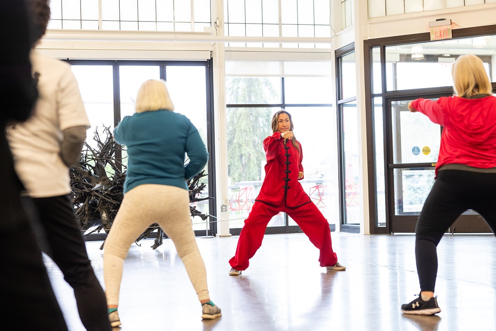 A woman dressed in red leads a standing tai chi exercise indoors.