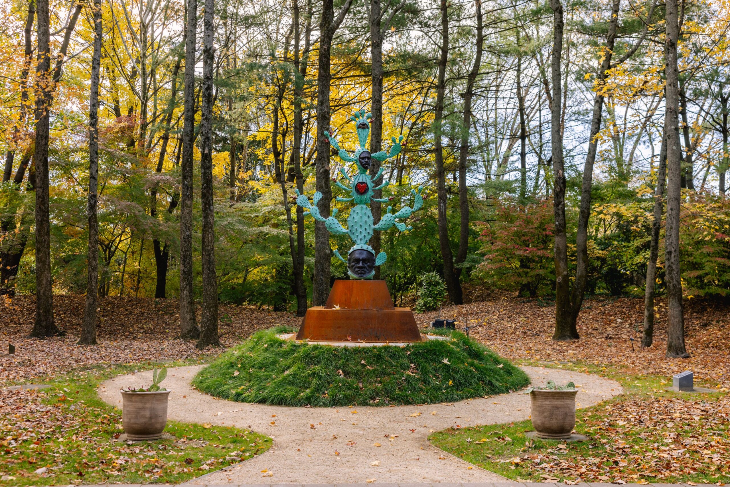 A tall green cactus sculpture with two heads built into it. It is outdoors on a bronze pedestal in a grove of trees.
