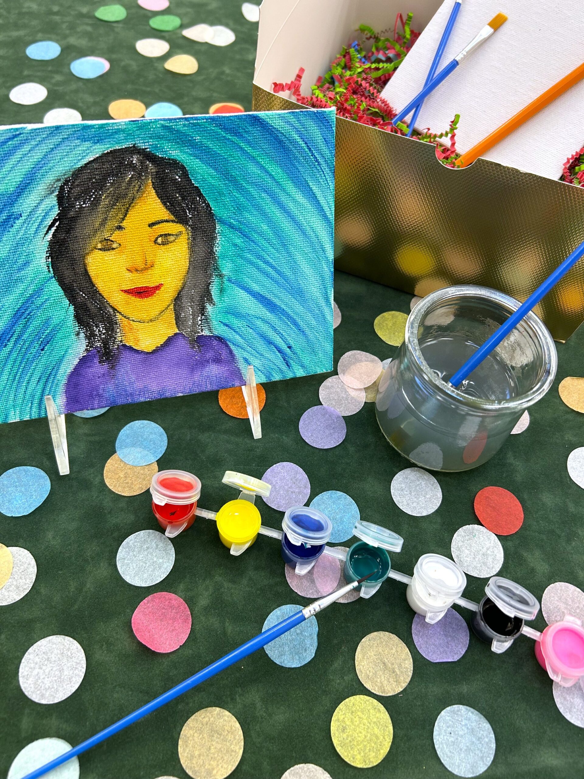A handpainted portrait of a woman in front of a set of paints and paintbrushes.
