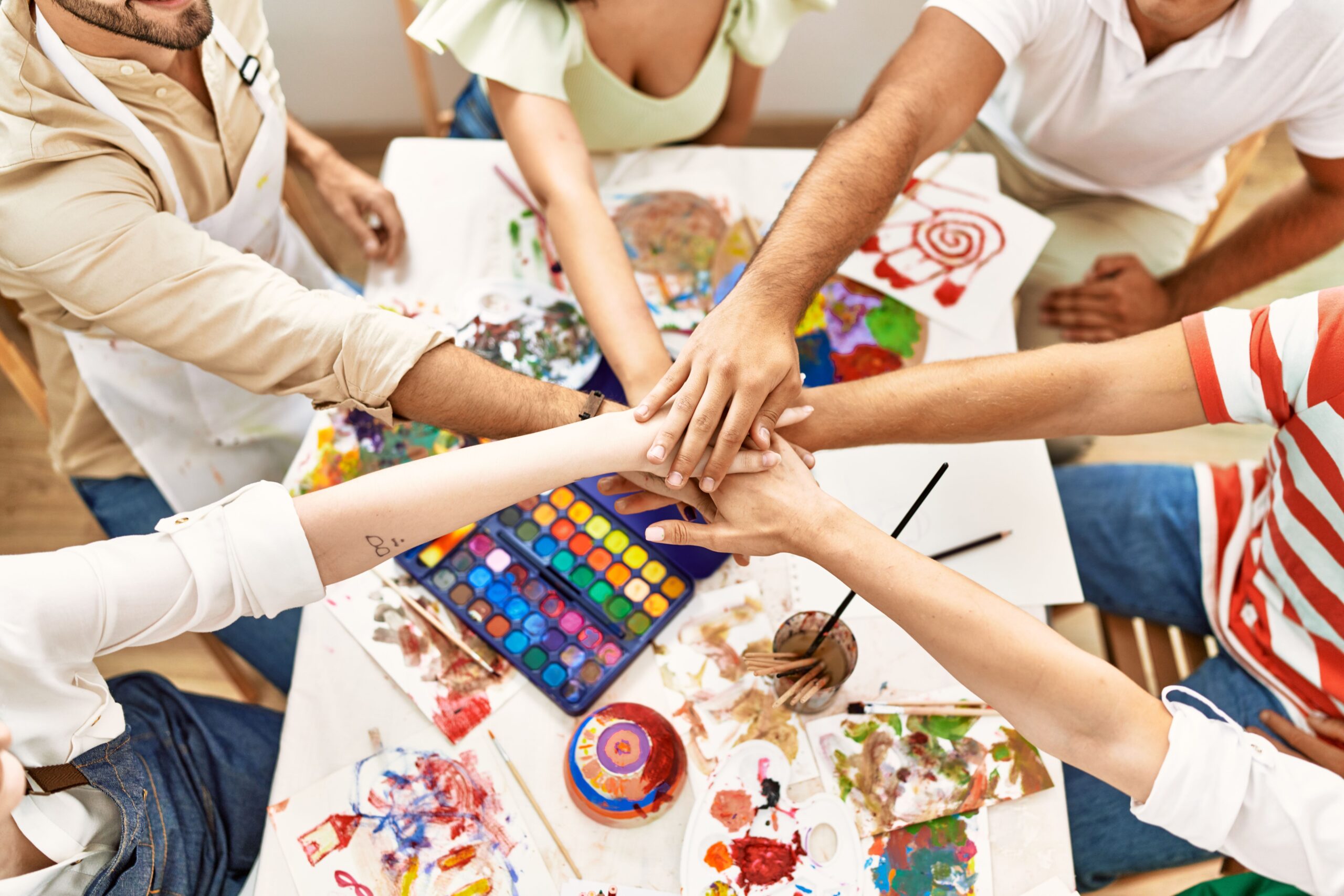 Six people in a circle holding their hands together over a table of art supplies.