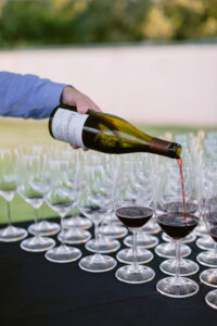A hand pouring red wine from a bottle into rows of wine glasses.