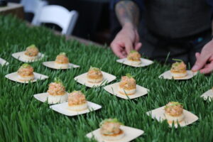 Hands arranging appetizers on wooden squares on a bed of fake grass.