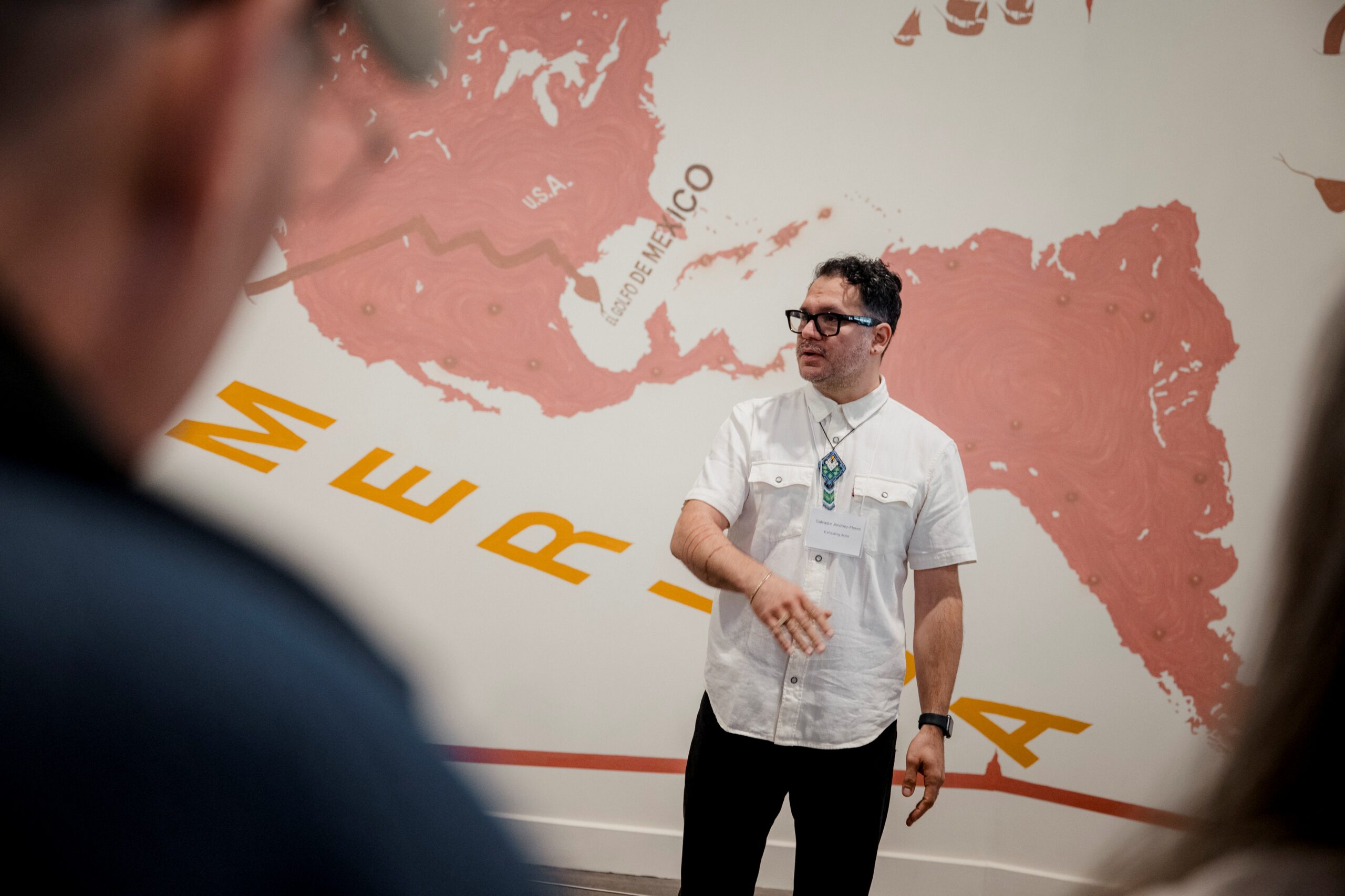 Artist Salvador Jiménez-Flores speaking indoors in front of the mural 'Memoria, Tierra, Trabajo: A Glimpse of the Semiquincentenial.'