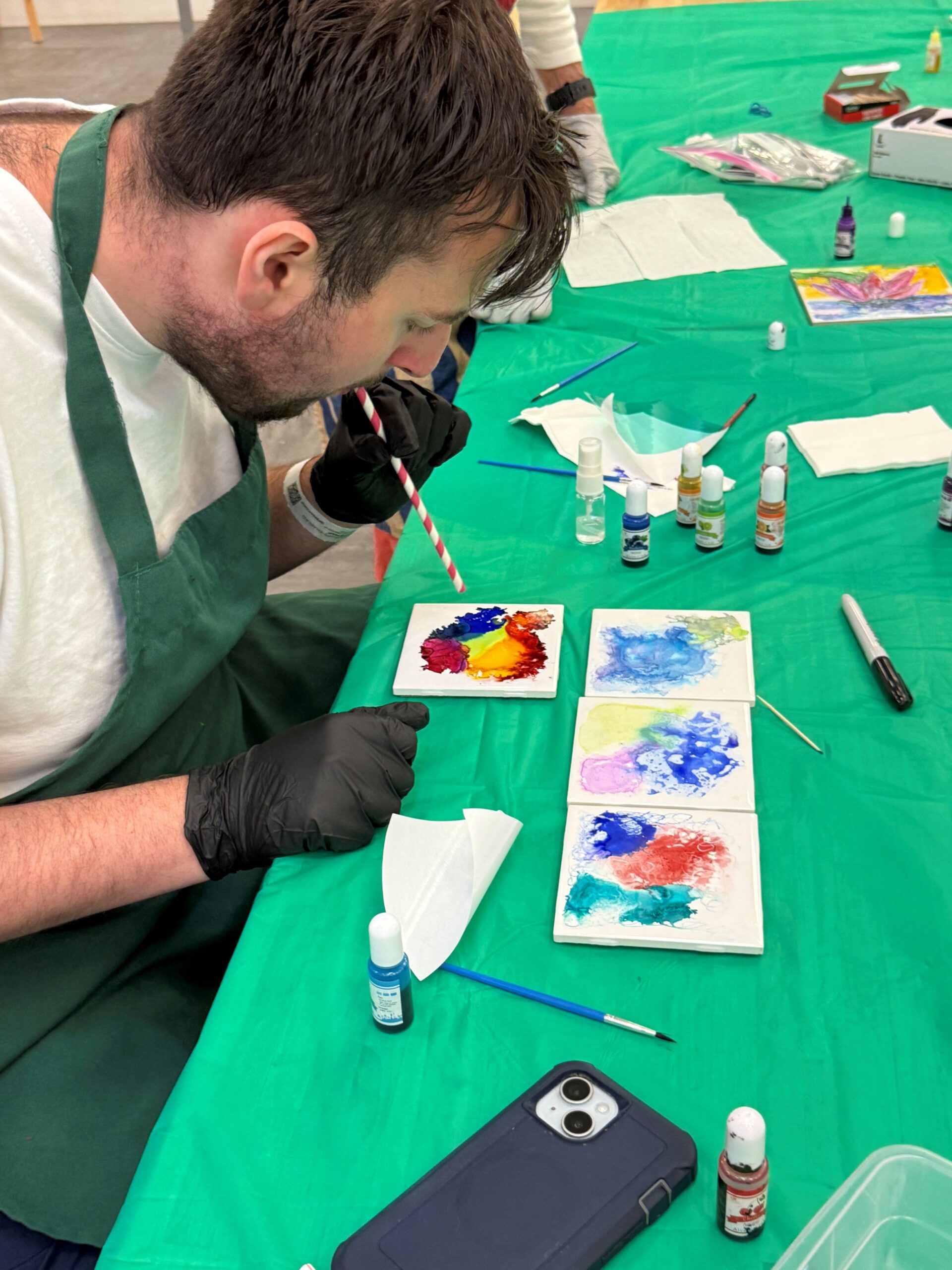 A man in a green apron seated at a table painting bright colors onto a white tile.