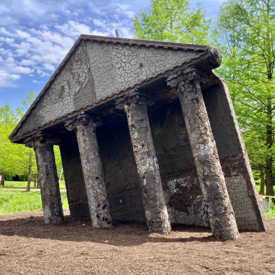 Row of tall, cracked stone columns forming a ruined temple exterior in a park, with green trees and a blue sky above.