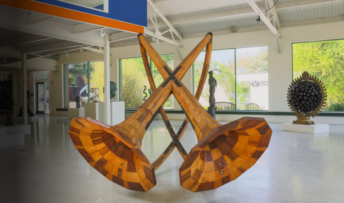 Two oversized wooden sculptures cross at the center to form an X, set in a bright museum gallery with large windows and other artworks in the background.