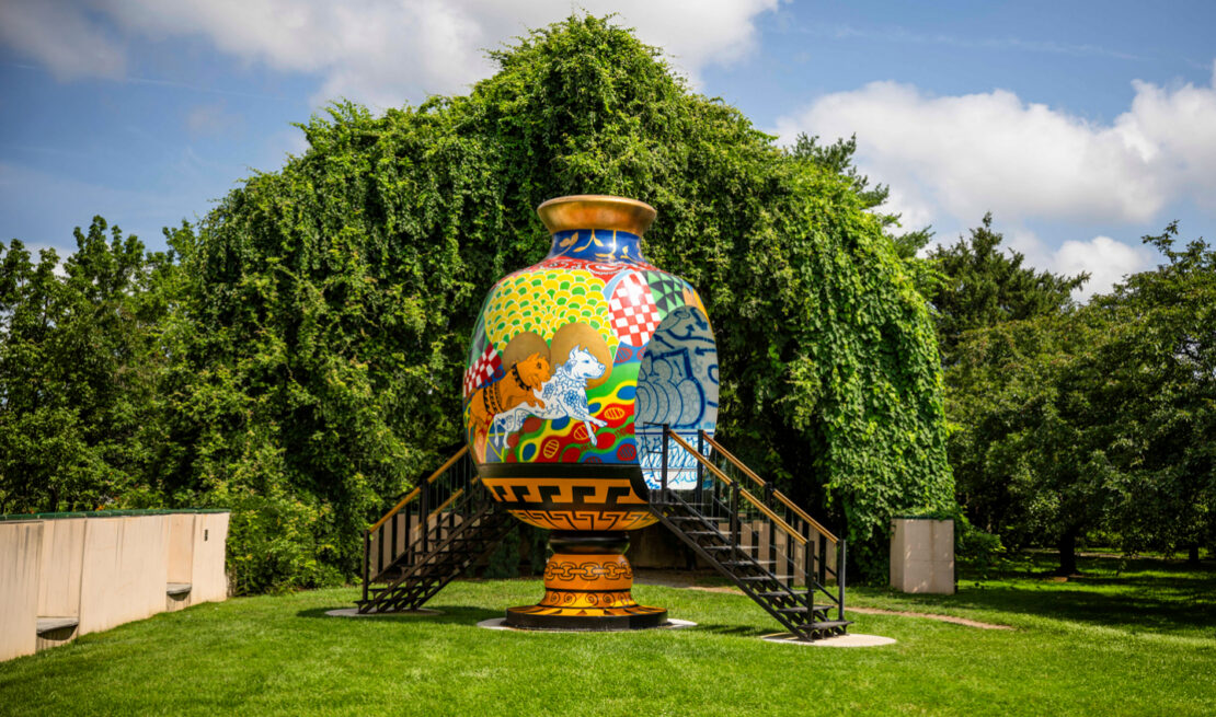 Colorful decorative vase sculpture with intricate patterns mounted on a pedestal, flanked by staircases in a sunlit park lawn.