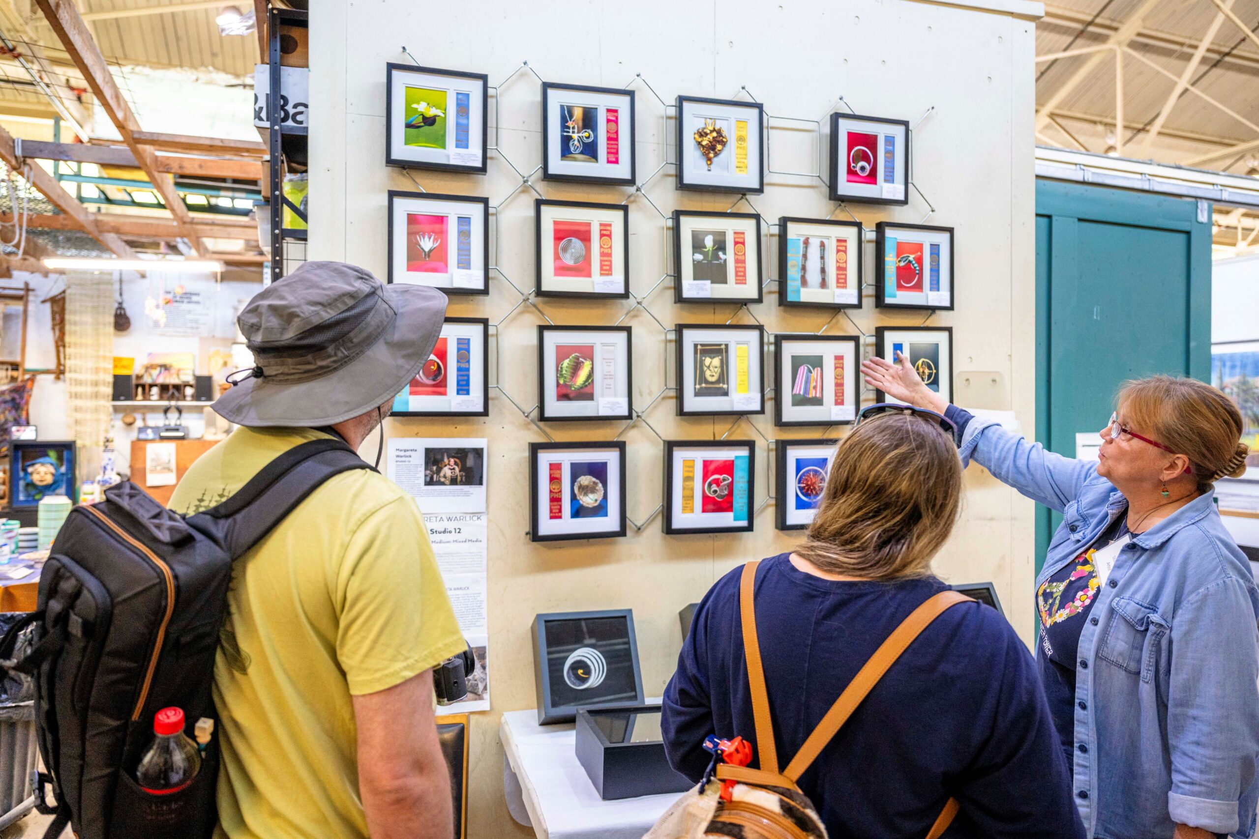 An artist speaks to two people and points to her framed work on the walls.