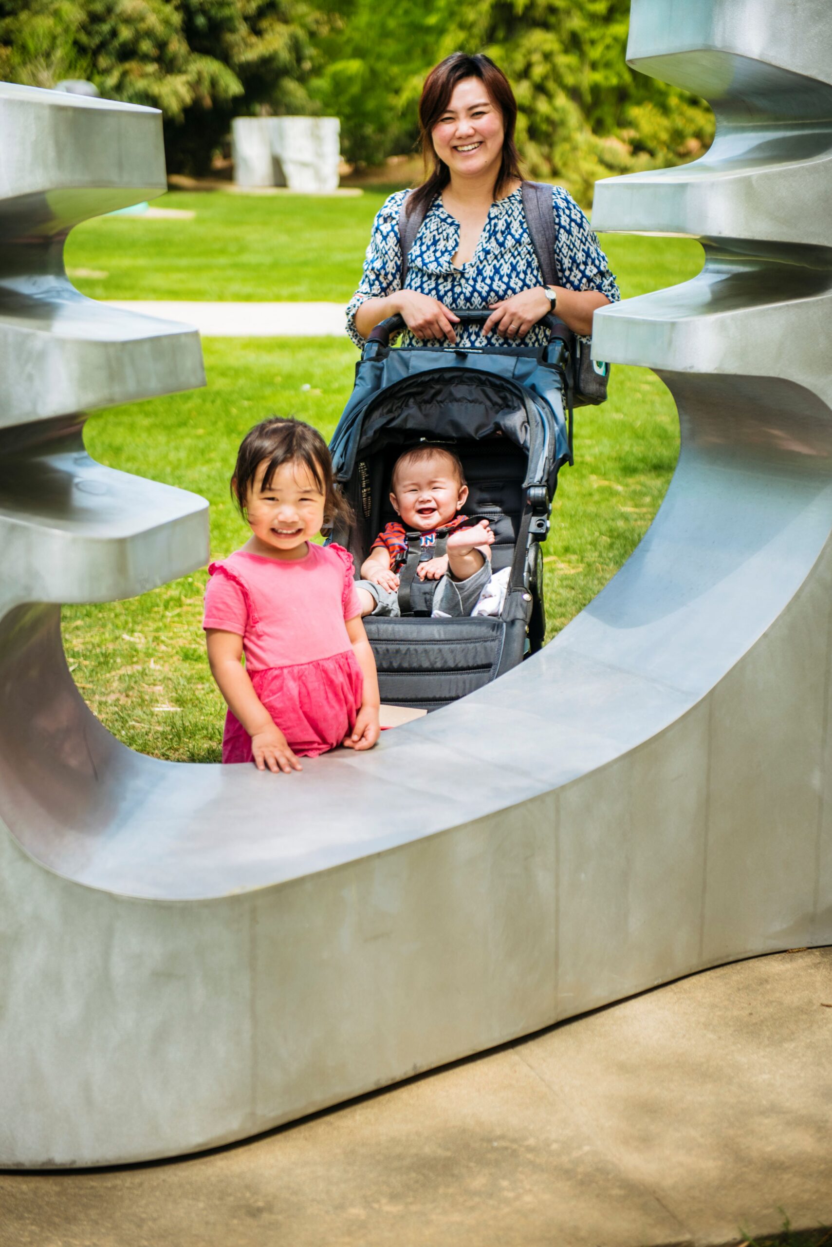 A woman and her two young children smile behind a sculpture. One of the children is in a stroller.