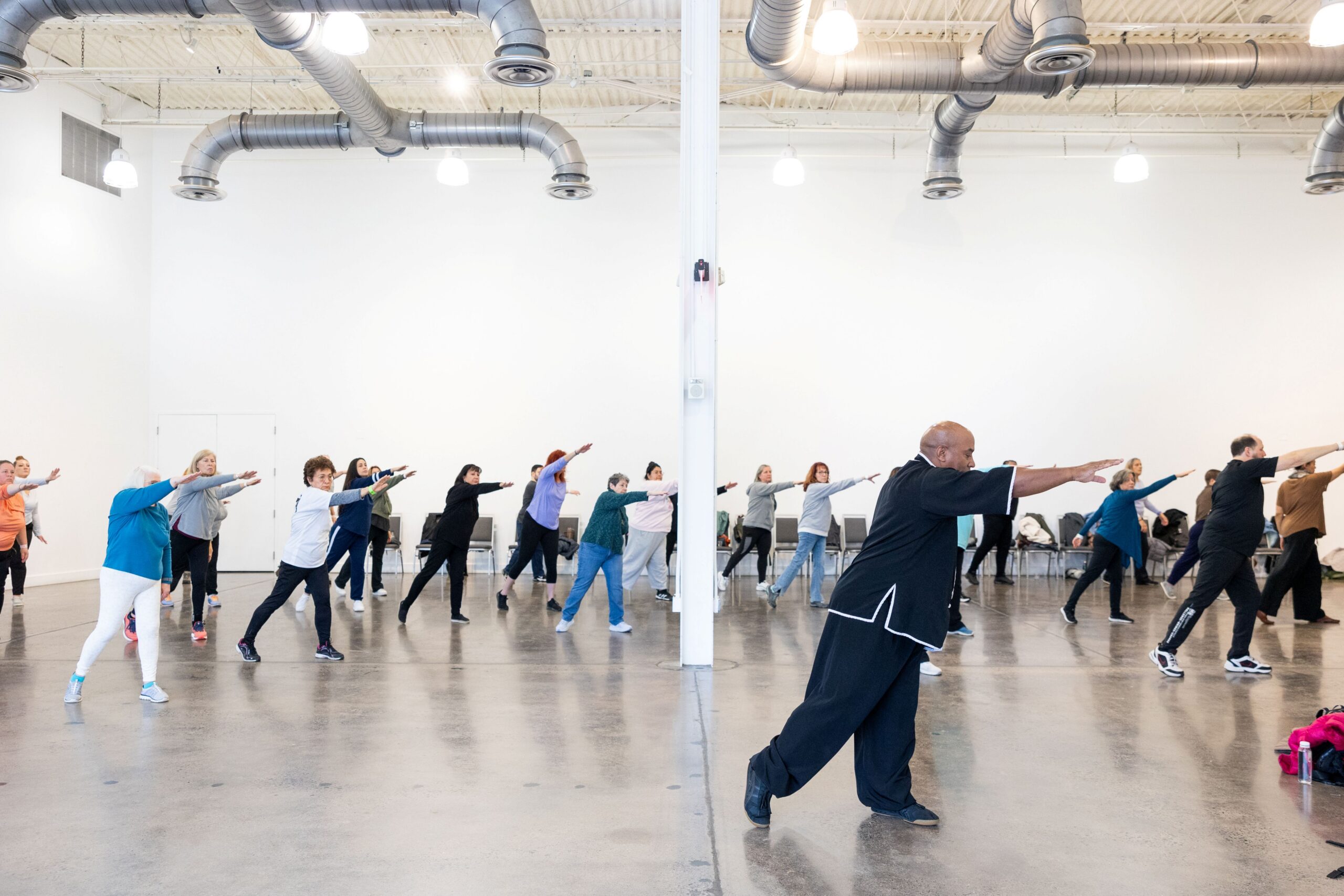 A man dressed in black leads a room full of people through a standing Tai Chi exercise.